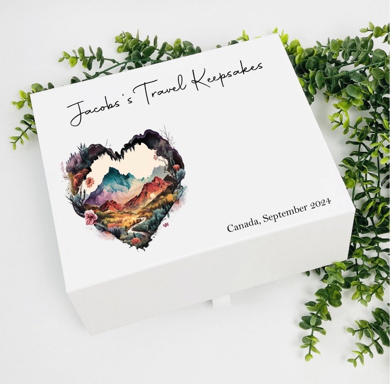 May include: White keepsake box with a heart-shaped design featuring a watercolor landscape of mountains and a winding road. The text "Jacob's Travel Keepsakes" and "Canada, September 2024" is printed on the box.