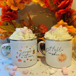 May include: Two white enamel mugs with silver rims, filled with whipped cream and marshmallows. One mug says "Cutest Pumpkin in the Patch" and the other says "Hello Pumpkin". Autumn-themed with fall foliage in the background.