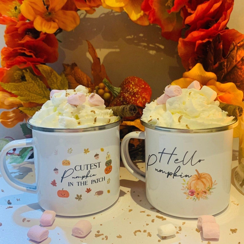 May include: Two white enamel mugs with silver rims, filled with whipped cream and marshmallows. One mug says "Cutest Pumpkin in the Patch" and the other says "Hello Pumpkin". Autumn-themed with fall foliage in the background.