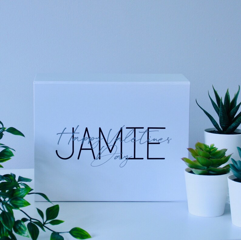 May include: White gift box with the text "Happy Valentine's Day" and the name "Jamie" in black lettering. The box is sitting on a white surface with two small potted succulents.