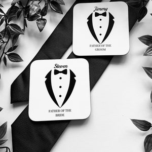 May include: Two white square coasters with black tuxedo designs and the text "Father of the Groom" and "Father of the Bride". The coasters have the names "Jimmy" and "Steven" printed on them.