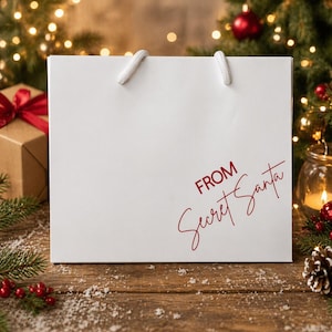 May include: A white gift bag with white rope handles. The words "FROM Secret Santa" are printed in red script on the front. The bag is on a wooden surface with Christmas decorations in the background.