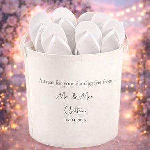Op de afbeelding: Een witte stoffen emmer gevuld met witte slippers. De emmer heeft twee handvatten en de tekst "A treat for your dancing feet from Mr & Mrs Cotton 17.04.2026". De achtergrond is een zacht, wazig bloemenontwerp.