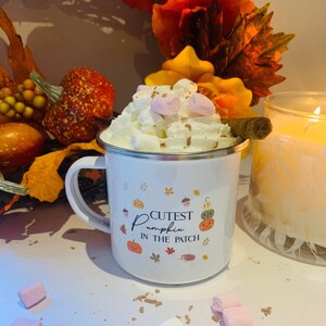 May include: White enamel mug with a silver rim, filled with whipped cream, marshmallows, and a cinnamon stick. The mug features a fall-themed design with pumpkins, acorns, and the text "Cutest Pumpkin in the Patch."