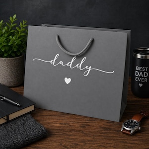 May include: A gray gift bag with the word "daddy" in white script and a white heart. The bag has a gray rope handle. A black tumbler with "BEST DAD EVER" is visible.