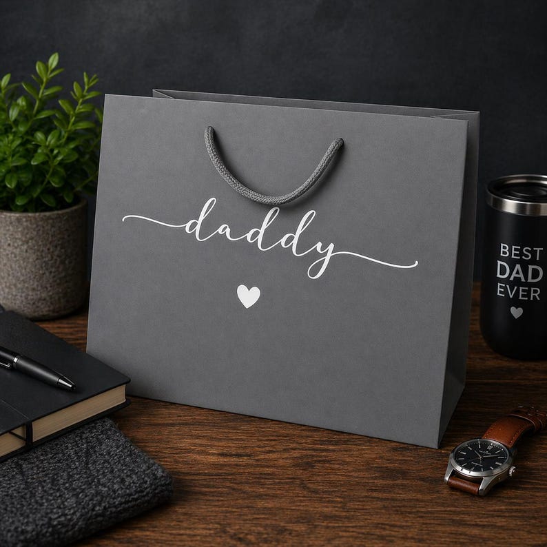 May include: A gray gift bag with the word "daddy" in white script and a white heart. The bag has a gray rope handle. A black tumbler with "BEST DAD EVER" is visible.