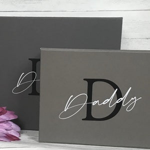 Pu&ograve; includere: Due cartelli in legno grigi con la lettera "D" in nero e la parola "Daddy" in corsivo bianco. I cartelli sono impilati uno sopra l'altro.