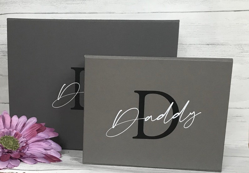 Pu&ograve; includere: Due cartelli in legno grigi con la lettera "D" in nero e la parola "Daddy" in corsivo bianco. I cartelli sono impilati uno sopra l'altro.