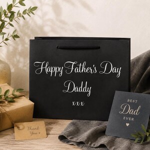 May include: A black gift bag with the text "Happy Father's Day Daddy" in white script. A small card that says "Best Dad Ever" is next to the bag. A wrapped gift and a "Thank You" tag are also visible.