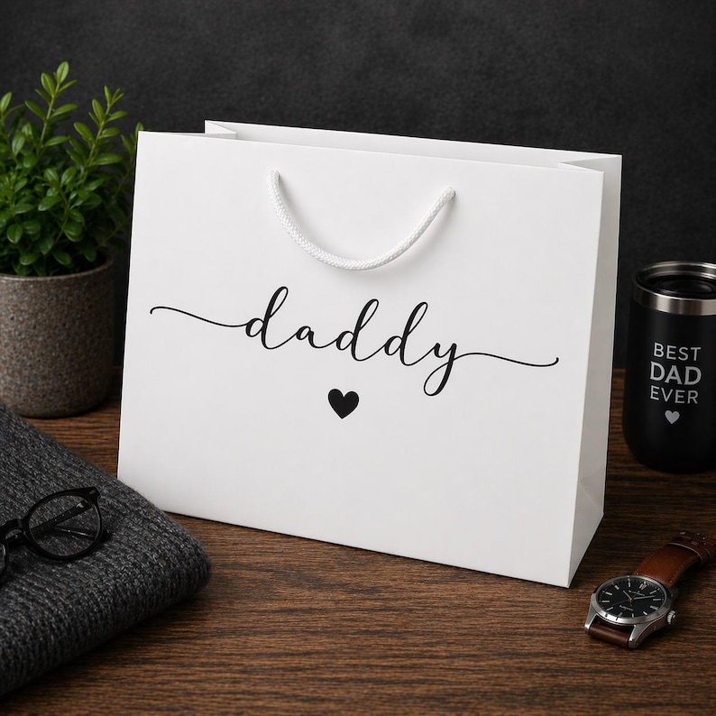 May include: A white gift bag with the word "daddy" in cursive script and a black heart. The bag has white rope handles. A black tumbler with "Best Dad Ever" is visible, along with a watch and a small plant.