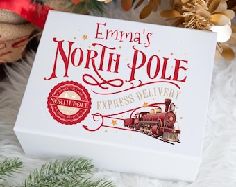 以下が含まれることがあります： 「Emma's North Pole Express Delivery」と赤い文字で書かれた白いギフトボックス。赤い列車のイラストと、「Santa Village North Pole Express Delivery」と書かれた円形のエンブレムが特徴です。
