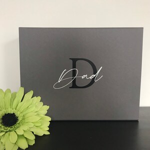 Pu&ograve; includere: Una scatola regalo grigia con una lettera nera "D" e la parola "Pap&agrave;" scritta in corsivo bianco.