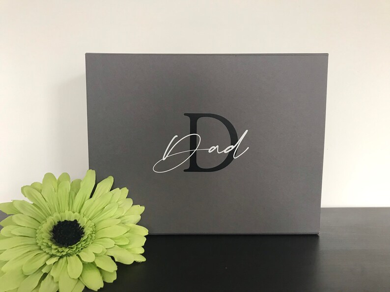 Pu&ograve; includere: Una scatola regalo grigia con una lettera nera "D" e la parola "Pap&agrave;" scritta in corsivo bianco.