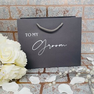 May include: A gray gift bag with a white rope handle. The bag has the text "To My Groom" printed on the front in white.