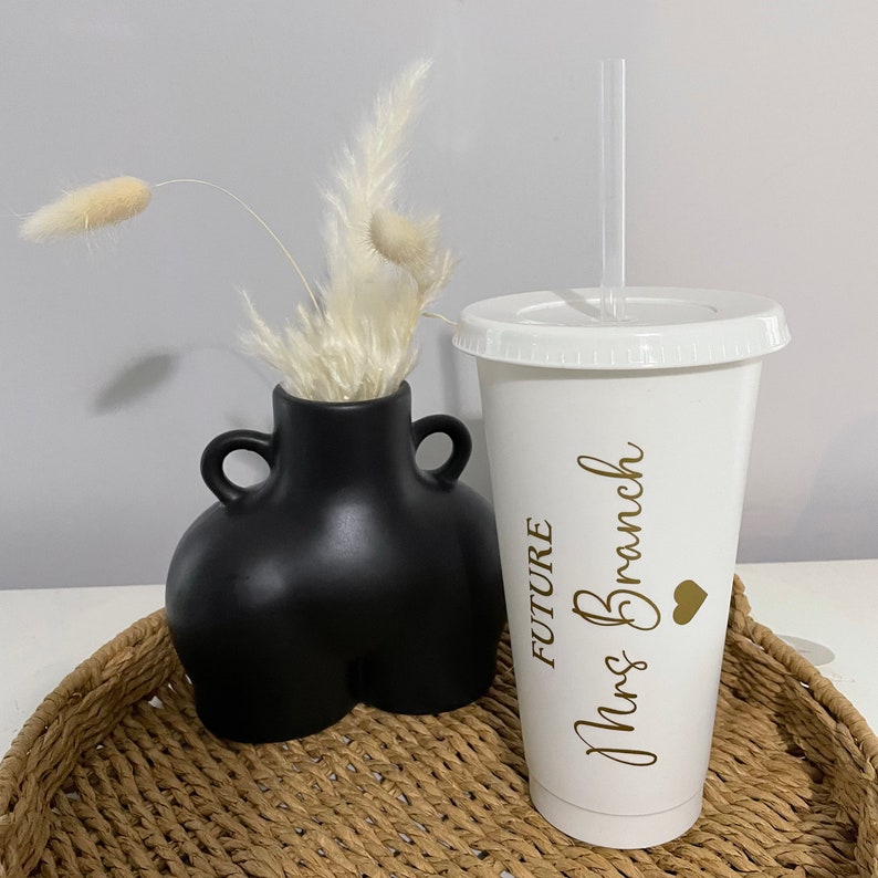 Puede incluir: Vaso reutilizable blanco con una pajita dorada y el texto "FUTURE Mrs. Branch" con un coraz&oacute;n dorado. El vaso est&aacute; sobre una bandeja de mimbre con un jarr&oacute;n negro con flores secas blancas.