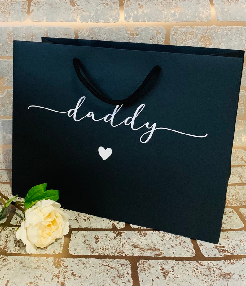 May include: A black gift bag with a white cursive "daddy" and a small white heart. The bag has black rope handles and a white flower is placed near the bottom left corner. The bag is on a brick background.