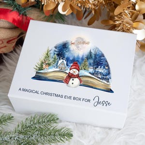 May include: White Christmas Eve box with a winter scene illustration. The box features a snowman, a train, and a house. Text on the box reads "A Magical Christmas Eve Box for Jesse."