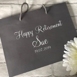 May include: Gray gift bag with white text that reads "Happy Retirement Sue 19.07.2019".