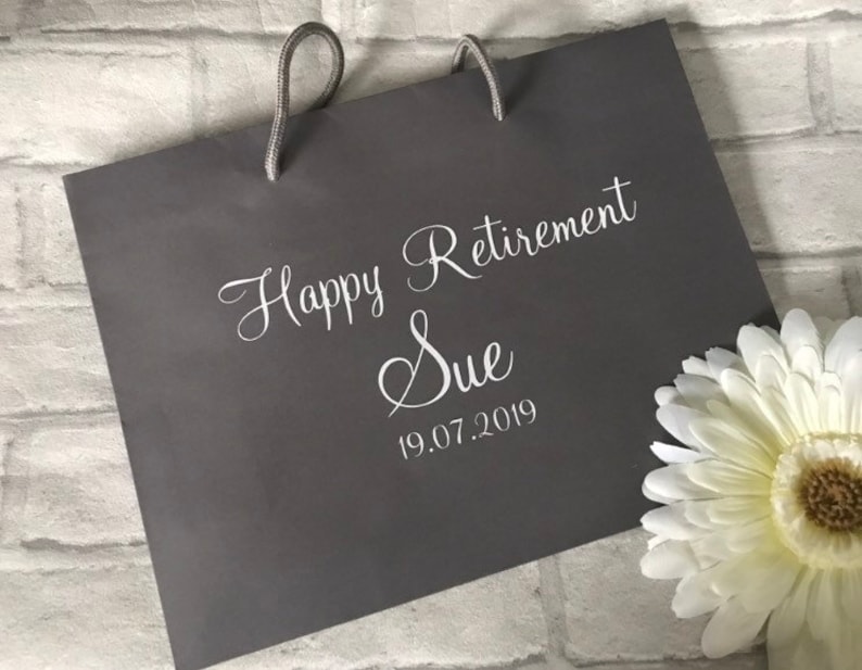 May include: Gray gift bag with white text that reads "Happy Retirement Sue 19.07.2019".