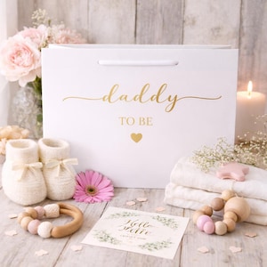 May include: A white gift bag with the words "daddy TO BE" in gold script, a pair of white baby booties, a pink flower, a wooden teether, and a card that says "Hello little one". A baby shower gift set.