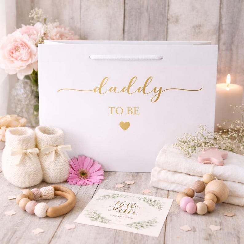 May include: A white gift bag with the words "daddy TO BE" in gold script, a pair of white baby booties, a pink flower, a wooden teether, and a card that says "Hello little one". A baby shower gift set.