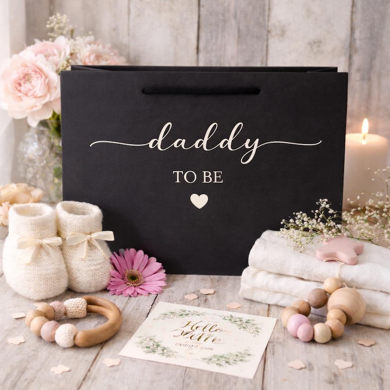 May include: A black gift bag with the words "daddy TO BE" in white script, a heart, and a pair of cream-colored baby booties. The scene includes a pink flower, a wooden teething ring, and a card.