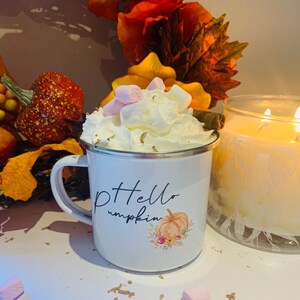 May include: A white enamel mug with a silver rim, filled with whipped cream, marshmallows, and a cinnamon stick. The mug has the text "Hello pumpkin" and a pumpkin graphic. A lit candle and fall foliage are in the background.