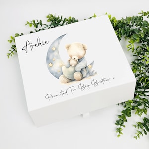 May include: White wooden box with a blue and white illustration of a teddy bear holding a smaller teddy bear on a crescent moon with stars. The text "Archie Promoted To Big Brother" is printed on the box.