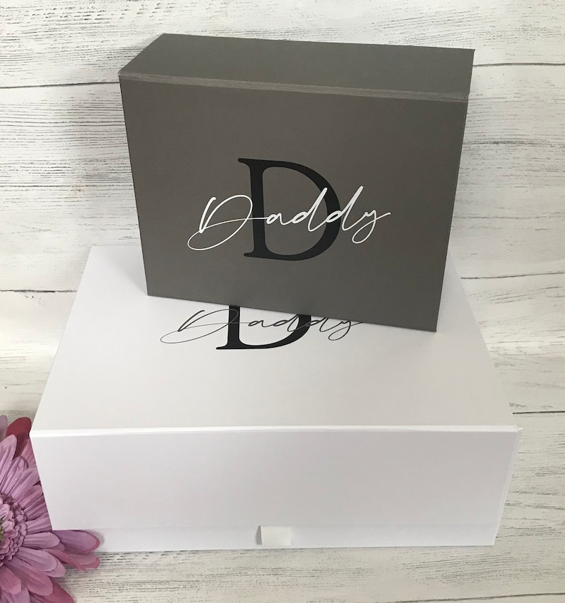 Pu&ograve; includere: Due scatole regalo impilate con un coperchio bianco e un coperchio grigio. Le scatole sono decorate con la lettera "D" e la parola "Daddy" in un font corsivo.