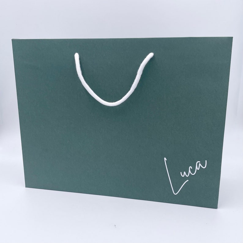 May include: A dark teal gift bag with a white rope handle. The word "Luca" is written in a cursive font on the lower right corner of the bag. The bag is rectangular and sits on a white surface.