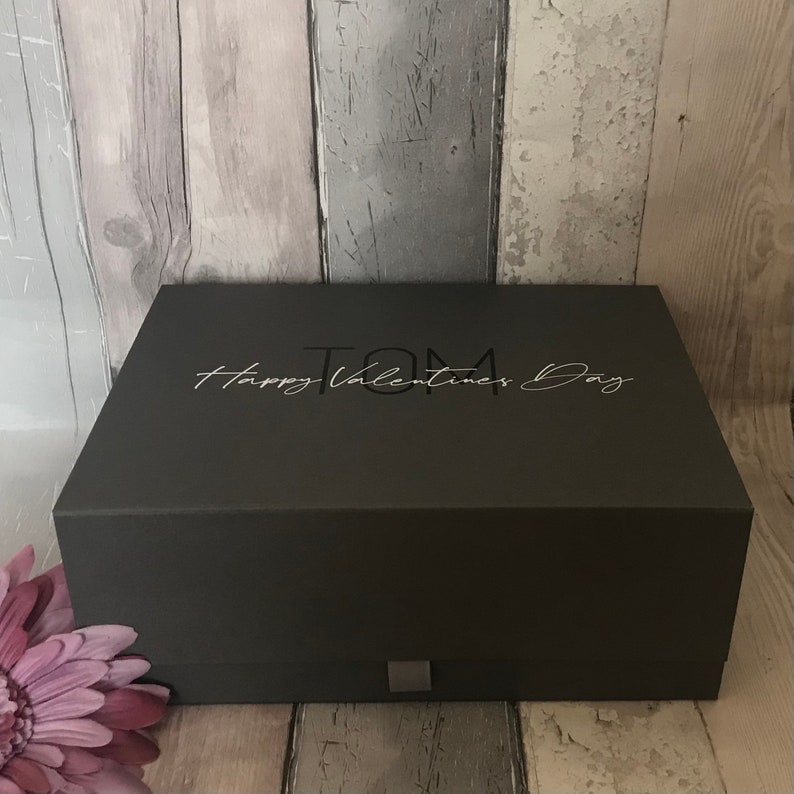 May include: A dark gray gift box with a silver inscription that reads "TOM Happy Valentines Day".