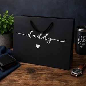 Puede incluir: Bolsa de regalo negra con la palabra "daddy" en escritura blanca y un pequeño corazón blanco. La bolsa tiene asas de cuerda negra. En el fondo se ve un vaso negro con "BEST DAD EVER", junto con un reloj y una cartera.