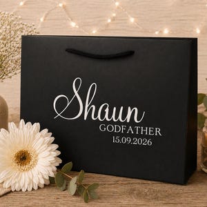 May include: A black gift bag with the name "Shaun" in elegant white script, along with "Godfather" and the date "15.09.2026". The bag has black rope handles. A white gerbera daisy and a lit candle are in the background.