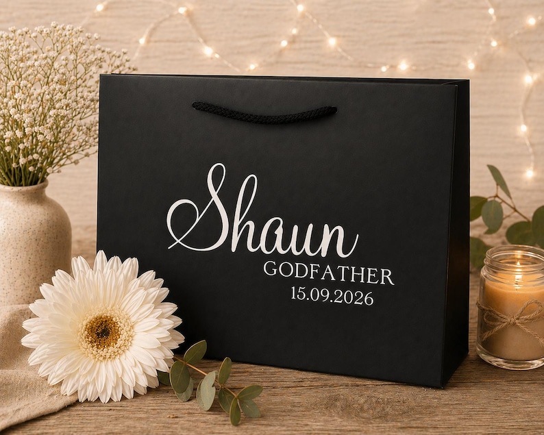 May include: A black gift bag with the name "Shaun" in elegant white script, along with "Godfather" and the date "15.09.2026". The bag has black rope handles. A white gerbera daisy and a lit candle are in the background.