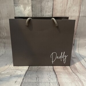 May include: A dark gray gift bag with rope handles. The word "Daddy" is written in a white script font on the front of the bag. The bag is sitting on a wooden surface.