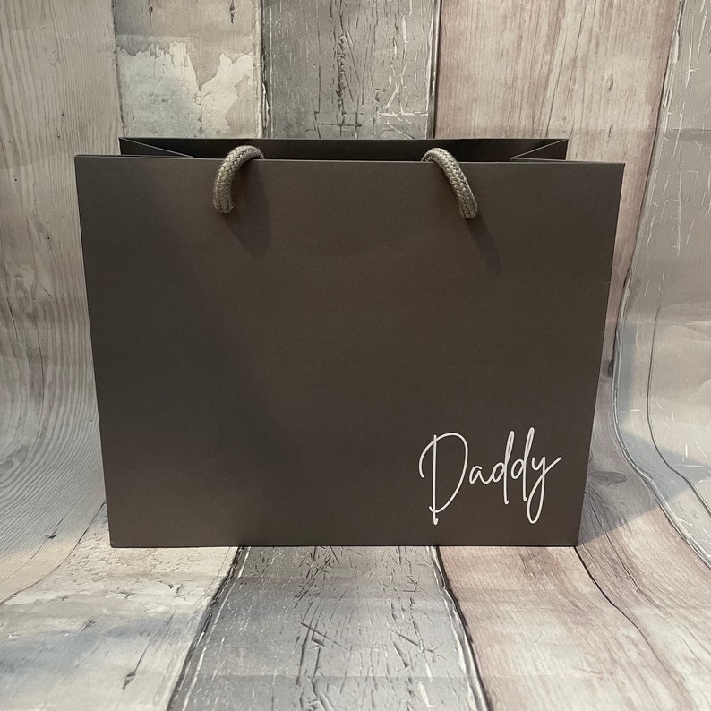 May include: A dark gray gift bag with rope handles. The word "Daddy" is written in a white script font on the front of the bag. The bag is sitting on a wooden surface.