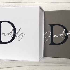 Pu&ograve; includere: Due scatole regalo con coperchi, una bianca e una grigia, entrambe con una lettera "D" nera e la parola "Daddy" scritta in grigio.