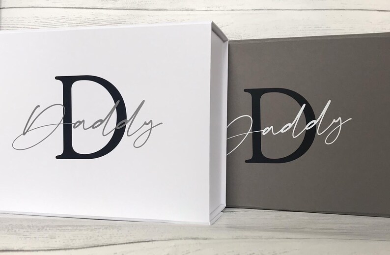 Pu&ograve; includere: Due scatole regalo con coperchi, una bianca e una grigia, entrambe con una lettera "D" nera e la parola "Daddy" scritta in grigio.