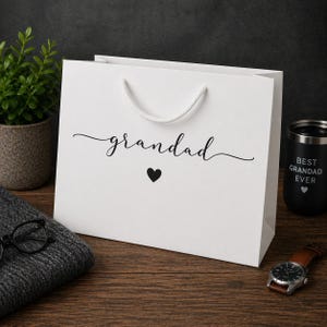 May include: A white gift bag with the word "grandad" in cursive script and a black heart. A black tumbler with "BEST GRANDAD EVER" is visible. A watch and glasses are also in the image.