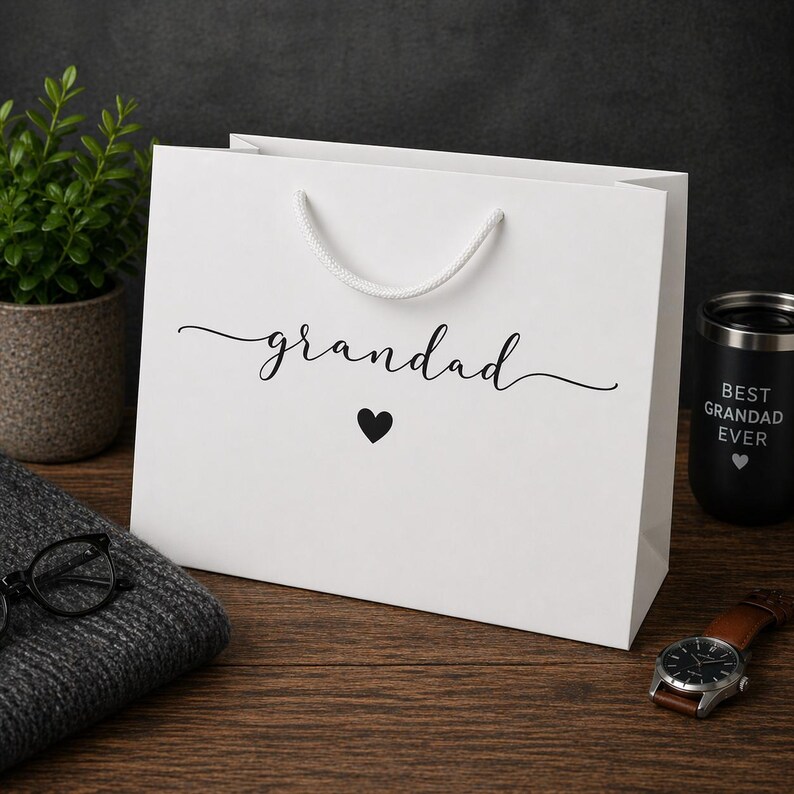 May include: A white gift bag with the word "grandad" in cursive script and a black heart. A black tumbler with "BEST GRANDAD EVER" is visible. A watch and glasses are also in the image.