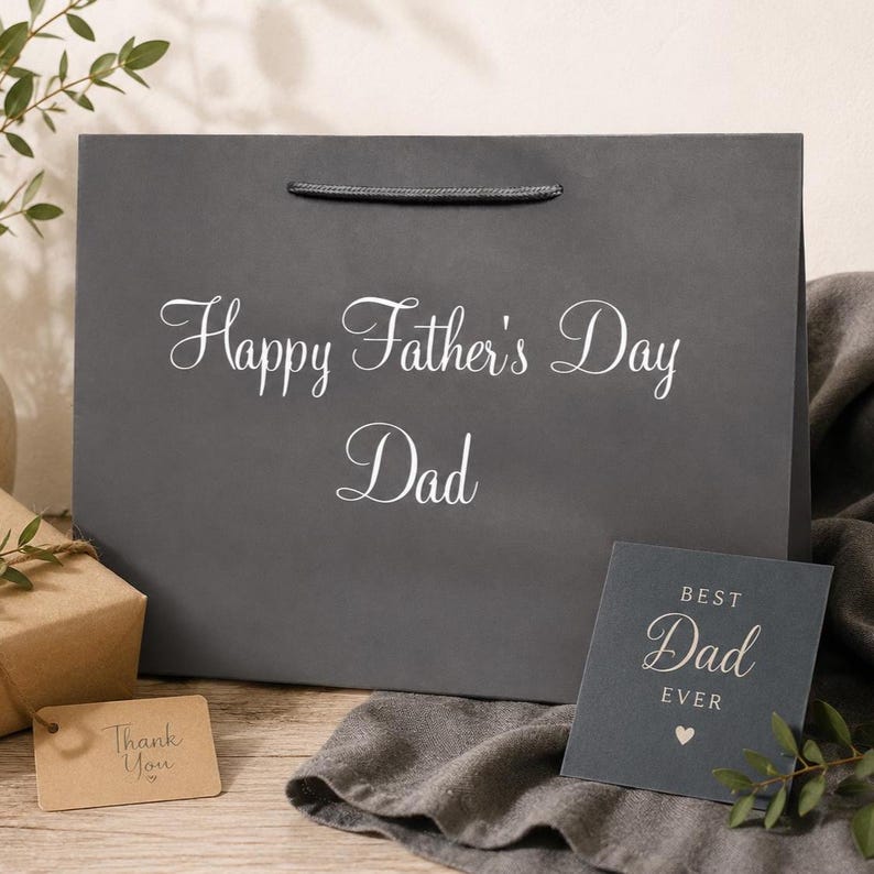 May include: A dark gray gift bag with the words "Happy Father's Day Dad" in white script. A small card next to the bag reads "Best Dad Ever" with a heart symbol. A small tag reads "Thank You".