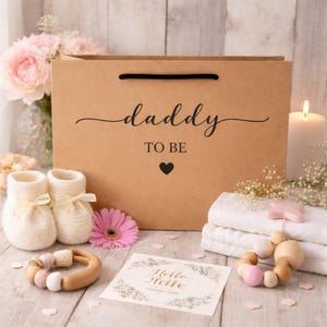 May include: A brown paper gift bag with the words "daddy TO BE" and a heart. The bag is surrounded by baby items, including white knit booties, a wooden teether, a greeting card, and a pink flower. A lit candle and flowers are in the background.