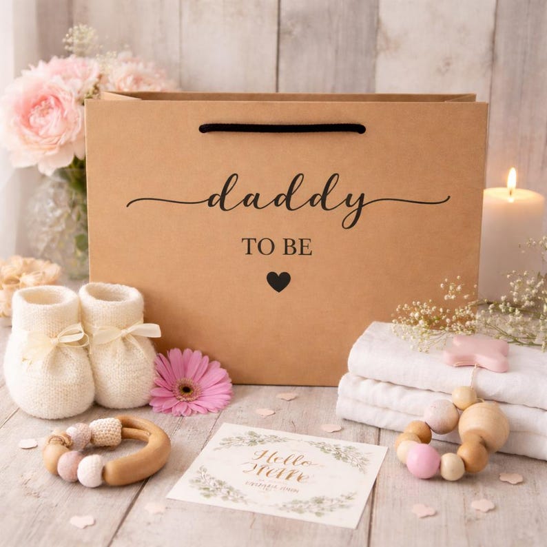 May include: A brown paper gift bag with the words "daddy TO BE" and a heart. The bag is surrounded by baby items, including white knit booties, a wooden teether, a greeting card, and a pink flower. A lit candle and flowers are in the background.