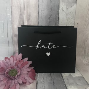 May include: A black gift bag with the name "Kate" in white script and a small white heart. The bag has black rope handles and is next to a pink flower. The background is a weathered wood.