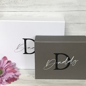 Pu&ograve; includere: Due scatole regalo, una bianca e una grigia. La scatola bianca ha una lettera "D" nera e il testo "Pap&agrave;" in corsivo. La scatola grigia ha una lettera "D" nera e il testo "Pap&agrave;" in corsivo.