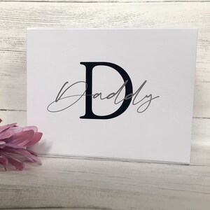 Pu&ograve; includere: Un biglietto bianco con una lettera nera "D" e la parola "Daddy" scritta in corsivo grigio.