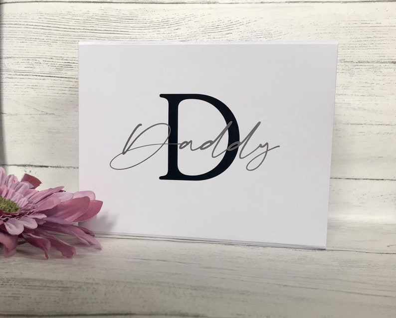 Pu&ograve; includere: Un biglietto bianco con una lettera nera "D" e la parola "Daddy" scritta in corsivo grigio.