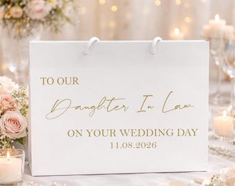 Puede incluir: Una bolsa de regalo blanca con texto dorado: "TO OUR DAUGHTER IN LAW ON YOUR WEDDING DAY 11.08.2026". La bolsa tiene asas de cuerda blanca. La escena sugiere una boda con flores y velas.