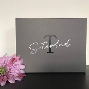 Pu&ograve; includere: Una scatola regalo grigia con una lettera "T" bianca e la parola "Stepdad" scritta in corsivo.