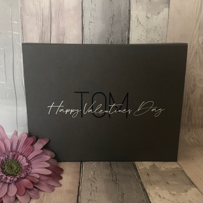 May include: A black gift box with the name "TOM" and the text "Happy Valentines Day" in silver lettering.